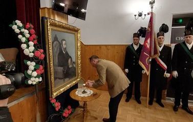 W&oacute;jt Grzegorz Węgrzynowski zapala symboliczną lampę Ignacego Łukasiewicza przy obrazie jubilata w sali Gminnego Centrum Kultury 