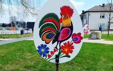 Metrowa pisanka wykonana z drewna. Na niej kolorowy kogucik. Pisanka umieszczona na skwerku w Chork&oacute;wce. 