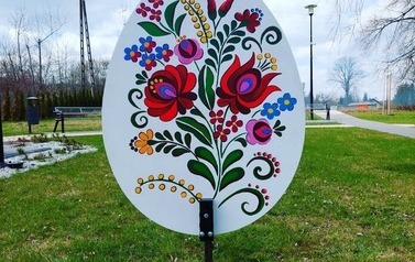 Metrowa pisanka wykonana z drewna. Na niej kolorowe kwiaty. Pisanka umieszczona na skwerku w Chork&oacute;wce. 