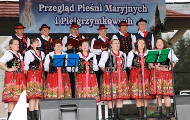 Zesp&oacute;ł śpiewaczy w ludowych strojach. Czerwone sp&oacute;dnice oraz gorsety.  Kobiety i mężczyźni. 
