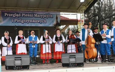 Zesp&oacute;ł śpiewaczy w ludowych strojach. Czerwone sp&oacute;dnice oraz gorsety.  Kobiety i mężczyźni. 