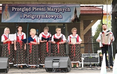 Zesp&oacute;ł śpiewaczy w ludowych strojach. ciemnozielone sp&oacute;dnice oraz gorsety.  Kobiety i mężczyźni. 