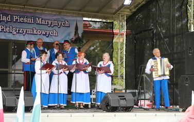 Zesp&oacute;ł śpiewaczy w ludowych strojach. jasnoniebieskie sp&oacute;dnice oraz gorsety.  Kobiety i mężczyźni. 