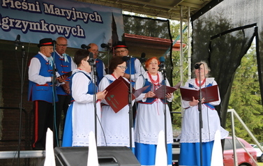 Zesp&oacute;ł śpiewaczy w ludowych strojach. jasnoniebieskie sp&oacute;dnice oraz gorsety.  Kobiety i mężczyźni. 