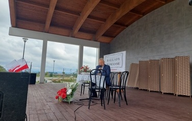 Narodowe czytanie w Gminie Chork&oacute;wka. Stacjonarna scena a na niej szyld Narodowego czytania, na białym tle czarne litery i napis Narodowe czytanie, wplecione elementy czerwone. Na scenie okrągły stolik na nim czerwony bukiet. Przy stoliku krzesła na kt&oacute;ry