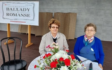 Narodowe czytanie w Gminie Chork&oacute;wka. Stacjonarna scena a na niej szyld Narodowego czytania, na białym tle czarne litery i napis Narodowe czytanie, wplecione elementy czerwone. Na scenie okrągły stolik na nim czerwony bukiet. Przy stoliku krzesła na kt&oacute;ry