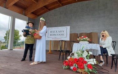 Narodowe czytanie w Gminie Chork&oacute;wka. Stacjonarna scena a na niej szyld Narodowego czytania, na białym tle czarne litery i napis Narodowe czytanie, wplecione elementy czerwone. Na scenie okrągły stolik na nim czerwony bukiet. Przy stoliku krzesła na kt&oacute;ry
