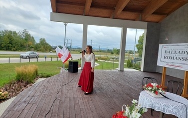 Narodowe czytanie w Gminie Chork&oacute;wka. Stacjonarna scena a na niej szyld Narodowego czytania, na białym tle czarne litery i napis Narodowe czytanie, wplecione elementy czerwone. Na scenie okrągły stolik na nim czerwony bukiet. Przy stoliku krzesła na kt&oacute;ry