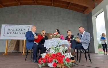 Narodowe czytanie w Gminie Chork&oacute;wka. Stacjonarna scena a na niej szyld Narodowego czytania, na białym tle czarne litery i napis Narodowe czytanie, wplecione elementy czerwone. Na scenie okrągły stolik na nim czerwony bukiet. Przy stoliku krzesła na kt&oacute;ry