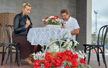 Narodowe czytanie w Gminie Chork&oacute;wka. Stacjonarna scena a na niej szyld Narodowego czytania, na białym tle czarne litery i napis Narodowe czytanie, wplecione elementy czerwone. Na scenie okrągły stolik na nim czerwony bukiet. Przy stoliku krzesła na kt&oacute;ry