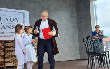 Narodowe czytanie w Gminie Chork&oacute;wka. Stacjonarna scena a na niej szyld Narodowego czytania, na białym tle czarne litery i napis Narodowe czytanie, wplecione elementy czerwone. Na scenie okrągły stolik na nim czerwony bukiet. Przy stoliku krzesła na kt&oacute;ry