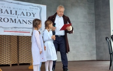 Narodowe czytanie w Gminie Chork&oacute;wka. Stacjonarna scena a na niej szyld Narodowego czytania, na białym tle czarne litery i napis Narodowe czytanie, wplecione elementy czerwone. Na scenie okrągły stolik na nim czerwony bukiet. Przy stoliku krzesła na kt&oacute;ry