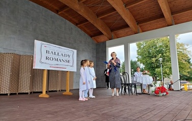 Narodowe czytanie w Gminie Chork&oacute;wka. Stacjonarna scena a na niej szyld Narodowego czytania, na białym tle czarne litery i napis Narodowe czytanie, wplecione elementy czerwone. Na scenie okrągły stolik na nim czerwony bukiet. Przy stoliku krzesła na kt&oacute;ry