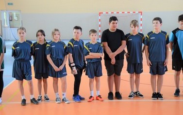 Drużyny szkolne rywalizujące w Gminnym Turnieju. Młodzież w kolorowych strojach sportowych. Młodzież stoi w rzędzie w budynku hali sportowej 