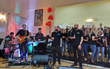Zdjęcie ch&oacute;ru mieszanego Legato. Ch&oacute;r w luźnych strojach dżinsach i koszulkach z nazwa ch&oacute;ry. w tych stylizacjach wykonywali swoje interpretacje piosenek wsp&oacute;łczesnych. Na przodzie solista ch&oacute;ru oraz dyrygentka. 