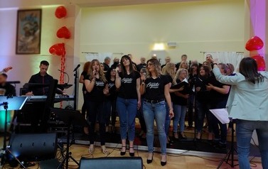 Zdjęcie ch&oacute;ru mieszanego Legato. Ch&oacute;r w luźnych strojach dzinsach i koszulkach z nazwa ch&oacute;ry. w tych stylizacjach wykonywali swoje interpretacje piosenek wsp&oacute;łczesnych. Na przodzie solista ch&oacute;ru oraz dyrygentka. 