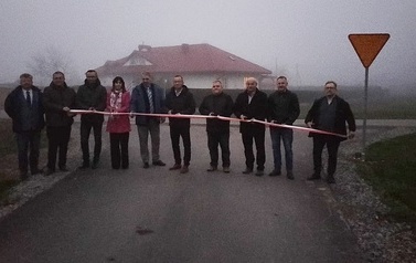 Otwarcie drogi. Grupa ludzi stoi w poprzek drogi przed szarfą, kt&oacute;ra ma być przecięta jako symbol oddania nowej drogi do użytku.   