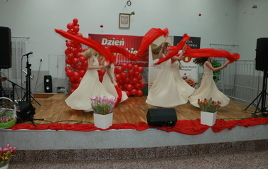 Dziewczyny w beżowych sukienkach trzymające w dłoniach czerwone wachlarze oraz czerwone zwiewne pasma materiał&oacute;w. Tanczą układ z wachlarzami. 