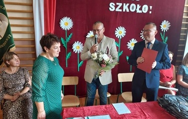 Na zdjęciu Helena Nowak oraz grono pedagogiczne. Są r&oacute;wnież W&oacute;jt i Zastępca kt&oacute;rzy wręczają kwiaty oraz dyplom. 