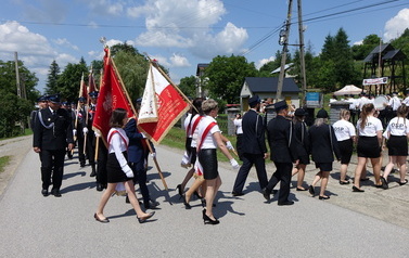 Zdjęcie wykonane podczas przemarszu uczestnik&oacute;w spotkania. Orkiestra Dęta na czele., tuz za nią strażacy w mundurach. Widoczne r&oacute;wnież poczty sztandarowe. 