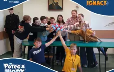Grupa dzieci trzymających wsp&oacute;lnie model cząsteczki wykonany z niebieskich i czerwonych kulek styropianowych połączonych wykałaczkami. Dzieci stoją w sali lekcyjnej, nad nimi widoczny jest portret mężczyzny. W tle stoi manekin w stroju g&oacute;rniczym.