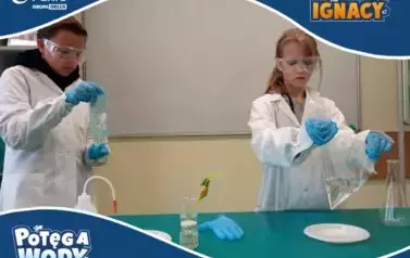 Na zdjęciu dwoje dzieci w białych fartuchach laboratoryjnych, goglach ochronnych i niebieskich rękawiczkach wykonuje eksperymenty chemiczne przy stole w klasie. Chłopiec po lewej stronie trzyma dwie połączone plastikowe butelki wypełnione wodą, tworząc ef