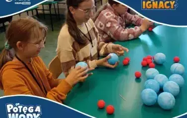 Grupa uczennic siedzi przy zielonym stole i wykonuje doświadczenie edukacyjne z niebieskimi i czerwonymi kulkami styropianowymi, prawdopodobnie tworząc modele cząsteczek. Dziewczynki są skupione na zadaniu. W tle widać klasę. Na grafice znajdują się loga 