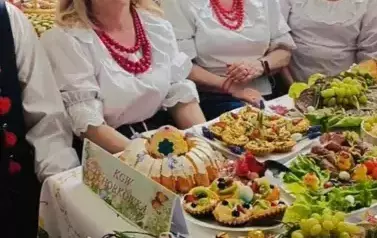 Grupa kobiet w tradycyjnych ludowych strojach siedzi przy bogato zastawionym stole, pełnym kolorowych dań wielkanocnych, ciast i dekoracji. Kobiety mają na sobie białe bluzki z koronkowymi kołnierzykami, ciemne gorsety i czerwone sp&oacute;dnice, a ich szyje zdo