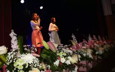 Na scenie występuje grupa dzieci i młodzieży ubranych w białe koszule i kolorowe sp&oacute;dnice (r&oacute;żowe i niebieskie). Śpiewają wsp&oacute;lnie, stojąc w kilku rzędach przed mikrofonami. Scena ozdobiona jest kompozycjami kwiatowymi w r&oacute;żowych doniczkach. Tło jest ciem