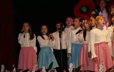 Grupa dorosłych os&oacute;b stoi razem na scenie, pozując do pamiątkowego zdjęcia. Większość z nich trzyma w rękach r&oacute;że. W tle czarna kurtyna i niebieskie światła sceniczne. Na pierwszym planie widoczne są kompozycje kwiatowe. Atmosfera wskazuje na oficjalne wy