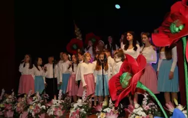 Grupa dorosłych os&oacute;b stoi razem na scenie, pozując do pamiątkowego zdjęcia. Większość z nich trzyma w rękach r&oacute;że. W tle czarna kurtyna i niebieskie światła sceniczne. Na pierwszym planie widoczne są kompozycje kwiatowe. Atmosfera wskazuje na oficjalne wy