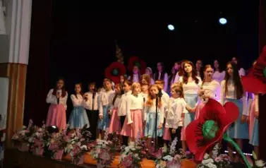 Grupa dorosłych os&oacute;b stoi razem na scenie, pozując do pamiątkowego zdjęcia. Większość z nich trzyma w rękach r&oacute;że. W tle czarna kurtyna i niebieskie światła sceniczne. Na pierwszym planie widoczne są kompozycje kwiatowe. Atmosfera wskazuje na oficjalne wy