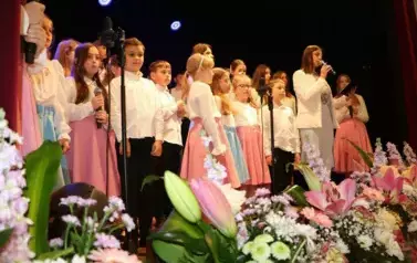 Grupa dorosłych os&oacute;b stoi razem na scenie, pozując do pamiątkowego zdjęcia. Większość z nich trzyma w rękach r&oacute;że. W tle czarna kurtyna i niebieskie światła sceniczne. Na pierwszym planie widoczne są kompozycje kwiatowe. Atmosfera wskazuje na oficjalne wy