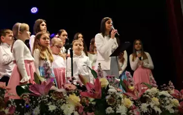 Grupa dorosłych os&oacute;b stoi razem na scenie, pozując do pamiątkowego zdjęcia. Większość z nich trzyma w rękach r&oacute;że. W tle czarna kurtyna i niebieskie światła sceniczne. Na pierwszym planie widoczne są kompozycje kwiatowe. Atmosfera wskazuje na oficjalne wy