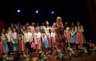 Grupa dorosłych os&oacute;b stoi razem na scenie, pozując do pamiątkowego zdjęcia. Większość z nich trzyma w rękach r&oacute;że. W tle czarna kurtyna i niebieskie światła sceniczne. Na pierwszym planie widoczne są kompozycje kwiatowe. Atmosfera wskazuje na oficjalne wy