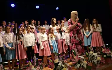 Grupa dorosłych os&oacute;b stoi razem na scenie, pozując do pamiątkowego zdjęcia. Większość z nich trzyma w rękach r&oacute;że. W tle czarna kurtyna i niebieskie światła sceniczne. Na pierwszym planie widoczne są kompozycje kwiatowe. Atmosfera wskazuje na oficjalne wy