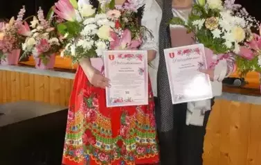 Pani Marysia w stroju ludowym obok niej Pani Teresa trzymają w dłoniach kwiaty. 