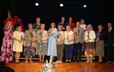Grupa dorosłych os&oacute;b stoi razem na scenie, pozując do pamiątkowego zdjęcia. Większość z nich trzyma w rękach r&oacute;że. W tle czarna kurtyna i niebieskie światła sceniczne. Na pierwszym planie widoczne są kompozycje kwiatowe. Atmosfera wskazuje na oficjalne wy
