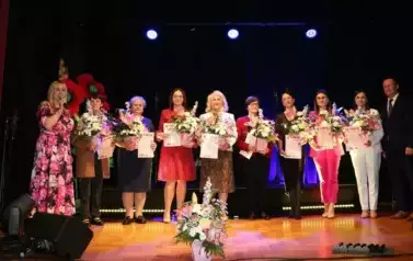 Grupa dorosłych os&oacute;b stoi razem na scenie, pozując do pamiątkowego zdjęcia. Większość z nich trzyma w rękach r&oacute;że. W tle czarna kurtyna i niebieskie światła sceniczne. Na pierwszym planie widoczne są kompozycje kwiatowe. Atmosfera wskazuje na oficjalne wy