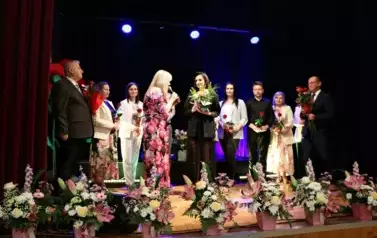 Grupa dorosłych os&oacute;b stoi razem na scenie, pozując do pamiątkowego zdjęcia. Większość z nich trzyma w rękach r&oacute;że. W tle czarna kurtyna i niebieskie światła sceniczne. Na pierwszym planie widoczne są kompozycje kwiatowe. Atmosfera wskazuje na oficjalne wy