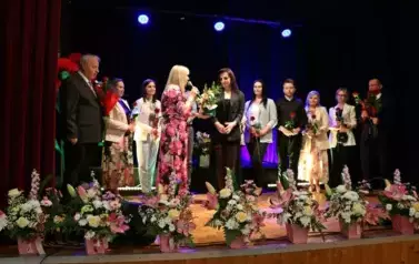 Grupa dorosłych os&oacute;b stoi razem na scenie, pozując do pamiątkowego zdjęcia. Większość z nich trzyma w rękach r&oacute;że. W tle czarna kurtyna i niebieskie światła sceniczne. Na pierwszym planie widoczne są kompozycje kwiatowe. Atmosfera wskazuje na oficjalne wy