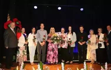 Grupa dorosłych os&oacute;b stoi razem na scenie, pozując do pamiątkowego zdjęcia. Większość z nich trzyma w rękach r&oacute;że. W tle czarna kurtyna i niebieskie światła sceniczne. Na pierwszym planie widoczne są kompozycje kwiatowe. Atmosfera wskazuje na oficjalne wy