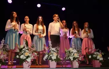 Na scenie występuje grupa dzieci i młodzieży ubranych w białe koszule i kolorowe sp&oacute;dnice (r&oacute;żowe i niebieskie). Śpiewają wsp&oacute;lnie, stojąc w kilku rzędach przed mikrofonami. Scena ozdobiona jest kompozycjami kwiatowymi w r&oacute;żowych doniczkach. Tło jest ciem