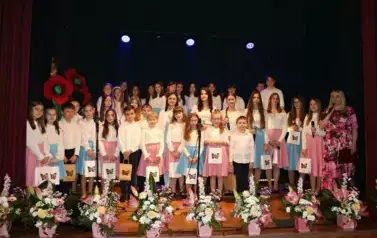 Na scenie występuje grupa dzieci i młodzieży ubranych w białe koszule i kolorowe sp&oacute;dnice (r&oacute;żowe i niebieskie). Śpiewają wsp&oacute;lnie, stojąc w kilku rzędach przed mikrofonami. Scena ozdobiona jest kompozycjami kwiatowymi w r&oacute;żowych doniczkach. Tło jest ciem