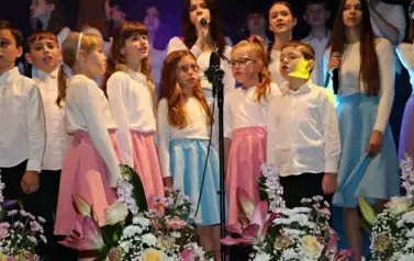 Na scenie występuje grupa dzieci i młodzieży ubranych w białe koszule i kolorowe sp&oacute;dnice (r&oacute;żowe i niebieskie). Śpiewają wsp&oacute;lnie, stojąc w kilku rzędach przed mikrofonami. Scena ozdobiona jest kompozycjami kwiatowymi w r&oacute;żowych doniczkach. Tło jest ciem