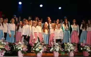 Na scenie występuje grupa dzieci i młodzieży ubranych w białe koszule i kolorowe sp&oacute;dnice (r&oacute;żowe i niebieskie). Śpiewają wsp&oacute;lnie, stojąc w kilku rzędach przed mikrofonami. Scena ozdobiona jest kompozycjami kwiatowymi w r&oacute;żowych doniczkach. Tło jest ciem