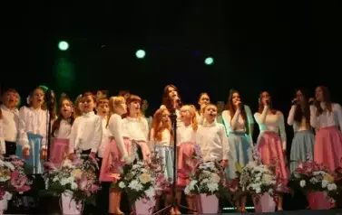 Na scenie występuje grupa dzieci i młodzieży ubranych w białe koszule i kolorowe sp&oacute;dnice (r&oacute;żowe i niebieskie). Śpiewają wsp&oacute;lnie, stojąc w kilku rzędach przed mikrofonami. Scena ozdobiona jest kompozycjami kwiatowymi w r&oacute;żowych doniczkach. Tło jest ciem