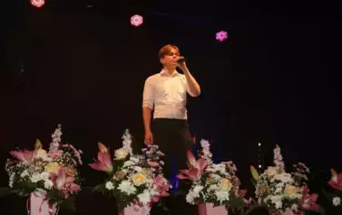 Chłopak w białej koszuli i ciemnych spodniach, śpiewa do mikrofonu. 