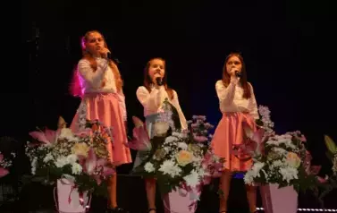 Na scenie występuje grupa dzieci i młodzieży ubranych w białe koszule i kolorowe sp&oacute;dnice (r&oacute;żowe i niebieskie). Śpiewają wsp&oacute;lnie, stojąc w kilku rzędach przed mikrofonami. Scena ozdobiona jest kompozycjami kwiatowymi w r&oacute;żowych doniczkach. Tło jest ciem