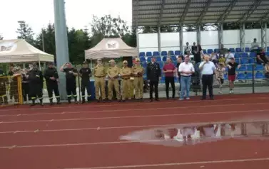 Grupa strażak&oacute;w w czarnych mundurach z odblaskowymi elementami stoi w zwartym szyku na zielonym boisku sportowym. W tle widać wysokie maszty oświetleniowe oraz drzewa i niebo z chmurami. Kilka os&oacute;b w środku grupy ubranych jest na czarno, ale bez odblask&oacute;w